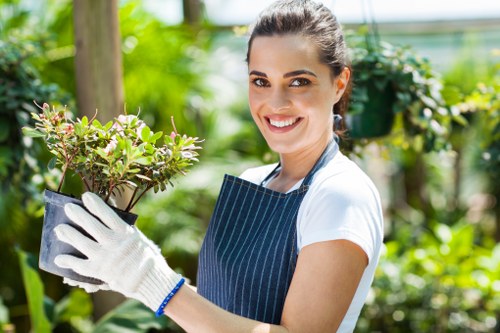 A woman with dark hair tied back, smiling and wearing a striped apron, white gloves, and a white t-shirt, holding a potted plant in an outdoor garden setting. The garden features a well-maintained lawn with dense grass, surrounded by various green plants, shrubs, and trees. In the background, there are tall trees with wooden trunks, and a green mesh or fence is visible. The lighting suggests it is a bright, sunny day, and the overall atmosphere reflects active gardening or landscaping work, consistent with outdoor maintenance services in the Rotherhithe area near London.