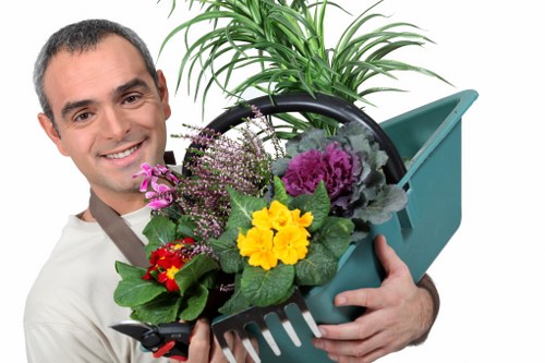 The image depicts a smiling man holding a green plastic garden watering can filled with a variety of flowering plants and green foliage. The plants include purple, yellow, and red flowers with lush green leaves, arranged in a way that showcases their vibrant colours. In the background, there is a neat lawn area with dense, healthy grass and a paved pathway made of grey stones. The garden environment appears well-maintained, with an outdoor setting featuring natural lighting and clear weather, suggesting a pleasant day for gardening. The scene emphasizes gardening activities such as planting and care, aligning with services like lawn maintenance and garden decoration offered by local providers in Rotherhithe, such as Hedge Trimming Rotherhithe, to help maintain attractive outdoor spaces.